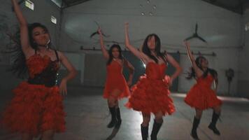 A group of dancers in red dresses performing in an industrial space. The lead dancer stands confidently in front, with others arranged behind her, all showcasing dynamic poses. video