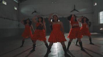 A group of young dancers performing in a dimly lit room, wearing vibrant red dresses. The energetic atmosphere is highlighted by their dynamic poses and movement video