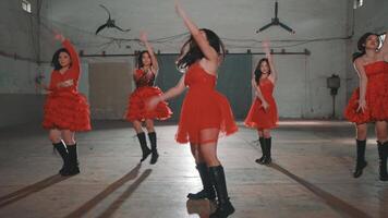 A group of dancers in red dresses performing energetically in an industrial space. The background features a spotlight and large fans, creating a dynamic atmosphere. video