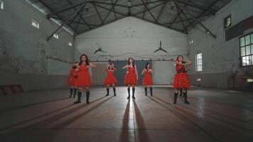 A group of dancers in red dresses performing in an industrial space with high ceilings and large windows. The floor reflects their shadows, creating a dramatic effect. video