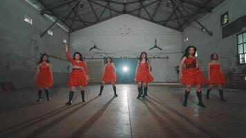 A group of dancers in red dresses performing in an industrial space. The dancers are synchronized, showcasing dynamic movements and expressions. The venue has a rustic video