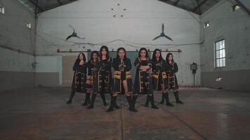 A group of six dancers performing in a spacious industrial setting. They wear matching black outfits with gold accents and strike powerful poses. The background features high ceilings and large fans. video