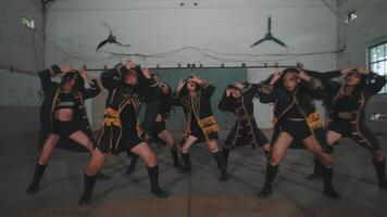A group of dancers performing energetically in a spacious, industrial-style room. They wear stylish black outfits with gold accents, showcasing synchronized movements and expressions of joy. video