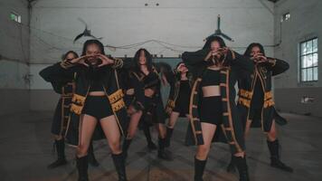 A group of dancers performing a synchronized routine in a spacious industrial setting. They wear black outfits with gold accents, showcasing dynamic movements and expressions. video