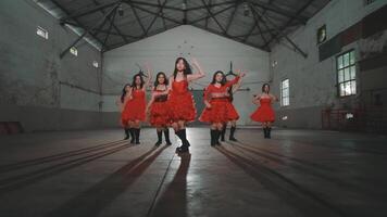 A group of young dancers performing in a spacious, industrial-style venue. They wear vibrant red dresses with ruffles and black boots, showcasing dynamic movements. The setting features high ceiling video