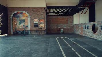 An empty indoor space featuring a brick wall with a colorful mural depicting a cafe scene. The floor is tiled with a checkerboard pattern, and there are a few chairs and tables arranged video