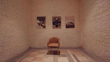 A minimalist gallery space featuring a single chair in the center. Three framed photographs hang on the white brick wall, showcasing various subjects. The floor has a patterned design, video