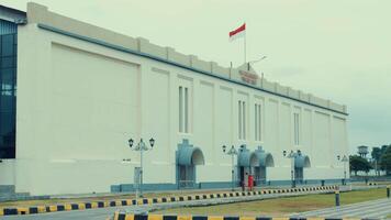 A large, white building with a flat roof and several arched entrances. The Indonesian flag is prominently displayed on top. The surrounding area features a paved road with yellow and black video