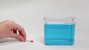 A hand places a matchstick in blue liquid, observing its buoyancy and movement in a clear container video