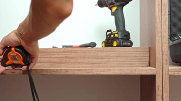 Close-up of man measuring wooden wardrobe with tape measure during furniture assembly, with tools and screws on shelf in background video