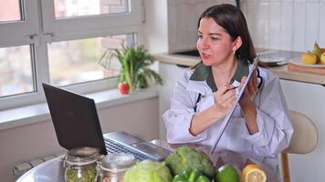 A nutritionist in a medical gown communicates online with patients at a table with fruits and vegetables, clearly showing a plan. Health and nutrition concept, diet, wellness. video