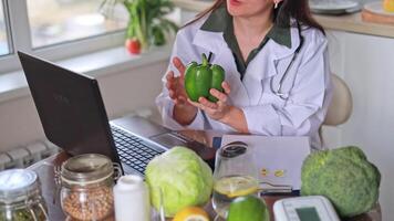 A nutritionist doctor in a medical gown communicates online with patients at a table with fruits and vegetables, clearly showing a green pepper. The concept of health and nutrition, diet, wellness. video