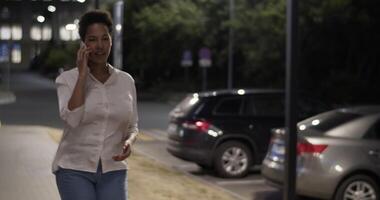 Ethnic businesswoman walking on street and speaking via mobile phone in evening video