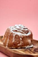 Cinnamon roll with icing on wooden platter against pink background photo