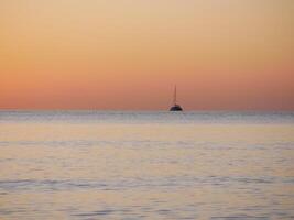 A breathtaking sunset over the calm sea with a sailboat on the horizon. Warm orange and golden tones fill the sky and reflect on the water. Travel, nature, relaxation, and maritime concept photo