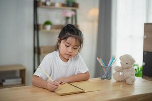 Young girl engaged in creative writing or drawing in a cozy indoor setting with soft light, surrounded by a teddy bear and colorful art supplies on a wooden table photo