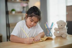 Young girl with long hair focused on writing on a notepad at home in a cozy and bright room with a teddy bear beside her on the table photo