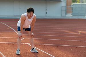 Asian man in white tank top looking ahead while standing with hand on knee, take a rest after run. photo
