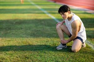 White tank top asian man with hands on his knee and ankle is crouching on grass field. Pain or injury. photo