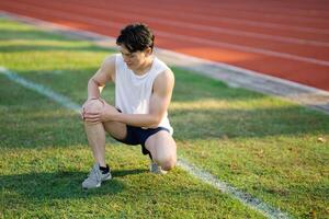 White tank top asian man is holding his knee while crouching on grass field. Pain or injury. photo