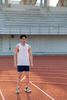 An asian man in white tank top is standing and looking ahead while taking a rest after running. photo