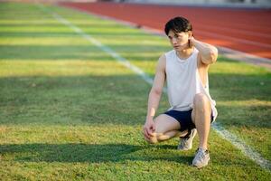 White tank top asian man with hands on his the neck is crouching on grass field. Waiting or resting. photo