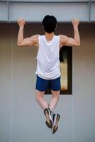 Full body of an asian man in white tank top doing a pull up on the horizontal bar in sports stadium. photo