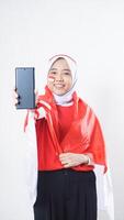 Indonesia independence day celebration with smiling Asian girl wearing red and white Indonesia flag color attribute showing blank screen smartphone to camera isolated on white background photo