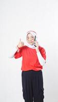 Indonesia independence day celebration with smiling Asian girl wearing red and white Indonesia flag color attribute talking on phone call while raising her thumb up isolated on white background photo