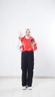 Indonesia independence day celebration with happy Asian girl wearing red and white Indonesia flag color attribute holding smartphone and showing OK sign with hand gesture isolated on white background photo