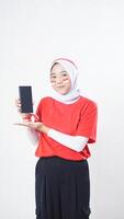 Indonesia independence day celebration with young adult Asian girl wearing red and white Indonesia flag color attribute showing smartphone blank screen to camera isolated on white background photo