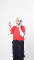 Indonesia independence day celebration with excited Asian girl wearing red and white Indonesia flag color attribute talking on phone call while raising her hand clenched isolated on white background photo