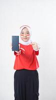 Indonesia independence day celebration with Asian girl wearing red and white Indonesia flag color attribute showing and pointing at smartphone with blank screen isolated on white background photo