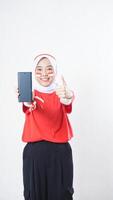 Indonesia independence day celebration with Asian girl wearing red and white Indonesia flag color attribute showing smartphone with blank screen and her thumb up isolated on white background photo