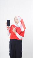 Indonesia independence day celebration with happy Asian girl wearing red and white Indonesia flag color attribute showing and pointing at blank screen smartphone to camera isolated on white background photo