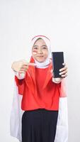Indonesia independence day celebration with Asian girl wearing red and white Indonesia flag color attribute showing and pointing at blank screen smartphone to camera isolated on white background photo