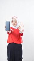 Indonesia independence day celebration with Asian girl wearing red and white Indonesia flag color attribute showing smartphone with blank screen and OK sign hand gesture isolated on white background photo