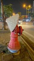 Fire hydrant dons conical hat at dusk, blending urban whimsy with East Asian folklore, echoing Lunar New Year celebrations photo
