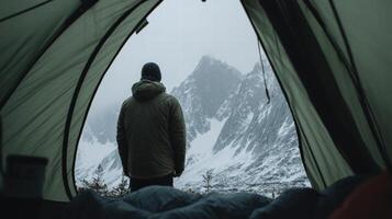 Man standing outside green tent looking at snowy mountain peak in cold weather photo