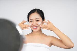Smiling cute Asian girl looking at the mirror, her hand is applying moisturizer lotion on her face photo