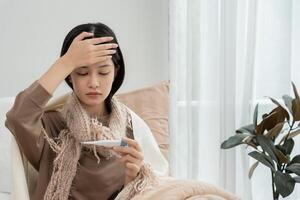 Young Asian woman having high fever while checking body temperature, female sneezing and runny nose with seasonal influenza, allergic, digital thermometer, virus, coronavirus, illness, photo