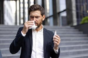 A businessman in a suit is blowing his nose outside, holding a nasal spray, likely suffering from allergies or a cold. photo