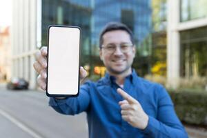 A man holds a phone with a blank screen, pointing at it in a business district. photo