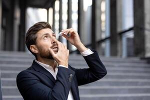 A businessman applying eye drops outdoors. He is wearing a suit and looking up with his mouth open. photo
