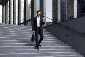 A businessman in a suit descends outdoor stairs while holding a phone and briefcase, modern city backdrop. photo
