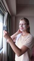 Woman with mask on the face looking out of the window in hotel room and using small mirror video