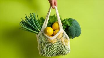 Hand holding mesh bag with fresh organic vegetables and fruits on green background photo