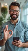 smiling man gestures with an okay sign while holding a smartphone. photo