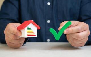 A man holds a green check mark near a house with an energy efficiency scale. Certificate of conformity to energy saving class. Green certification, utility cost savings, and eco-living. photo