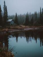 Serene Cabin Reflection in a Misty Forest Lake. A tranquil scene of a rustic cabin nestled amongst evergreens, perfectly mirrored in the still waters of a lake. photo